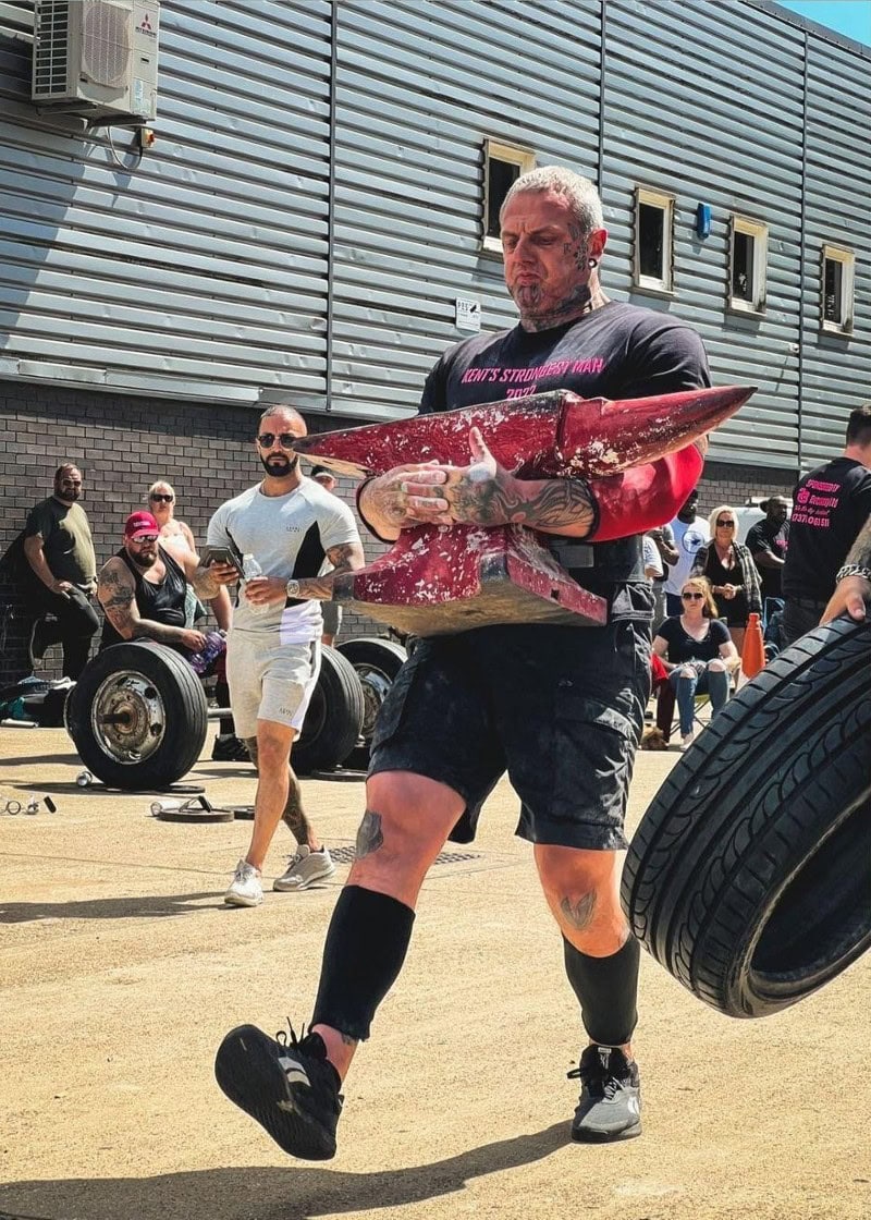 Paul Wood competing in a strongman event, demonstrating strength and technique while carrying a heavy anvil.